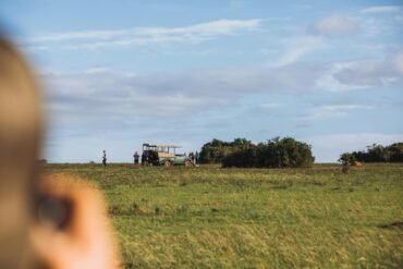 Unrecognizable person shooting picturesque landscape of grassy savanna and tourists standing near parked safari automobile on summer sunny day